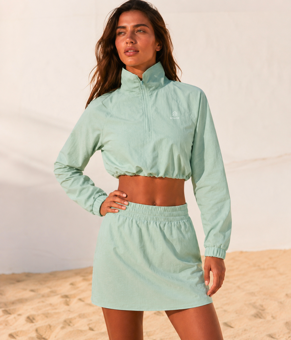 Model wearing the SCULVE sage quarter-zip and matching skirt on a sunlit beach